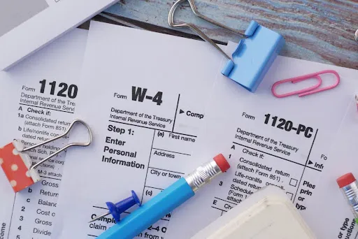 A close-up of W-4 and 1104 tax forms with pencils, paper clips, and binders on a table.