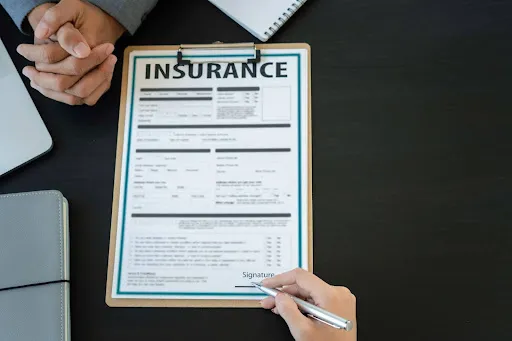 A clipboard displaying an insurance form is placed on a desk while a person prepares to sign it.