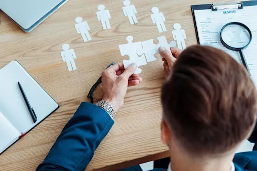 A top view of a man holding jigsaw puzzle pieces over a table with cutouts of people, a magnifying glass over a resume, a pen, notebook, and laptop, symbolizing human resources and recruitment strategies.