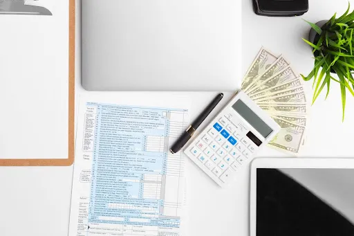 Tax form next to a clipboard, computer, calculator, money, pen, plant, and tablet.