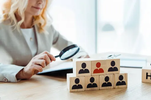 A blonde recruiter with a magnifying glass and a document examines wooden cubes with human icons, symbolizing human resources recruitment and evaluation.