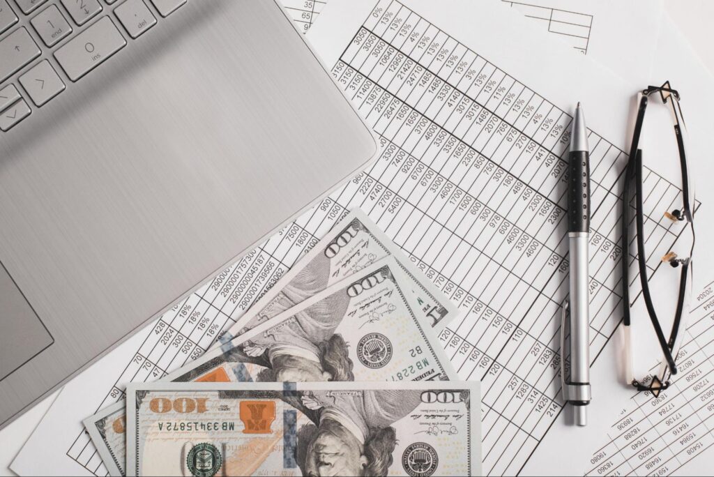 Top view of a desk with money, documents, a laptop, and a pen to be used in computing payroll.