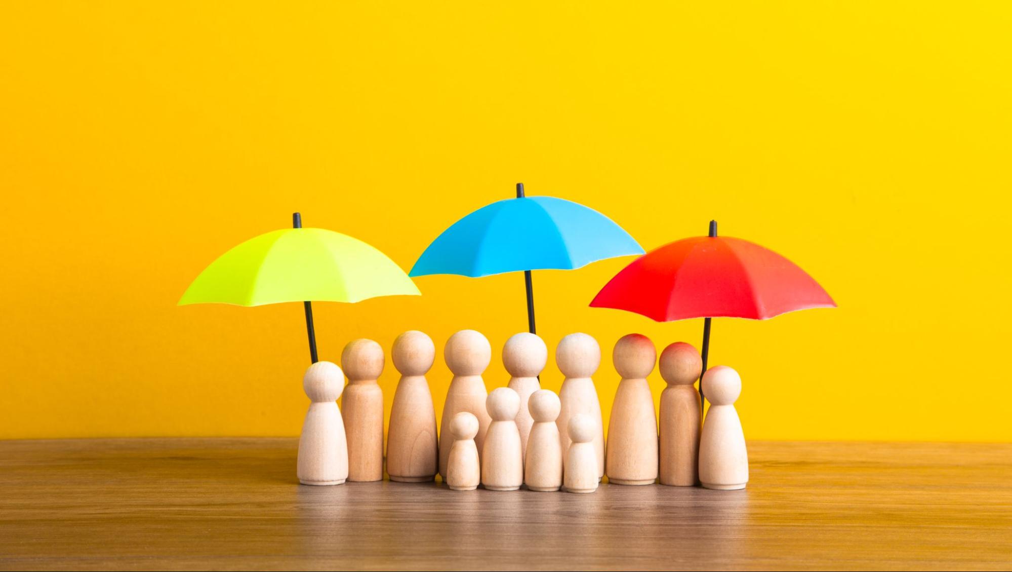 Wooden figures representing people protected by colorful umbrellas symbolizing liability insurance coverage.