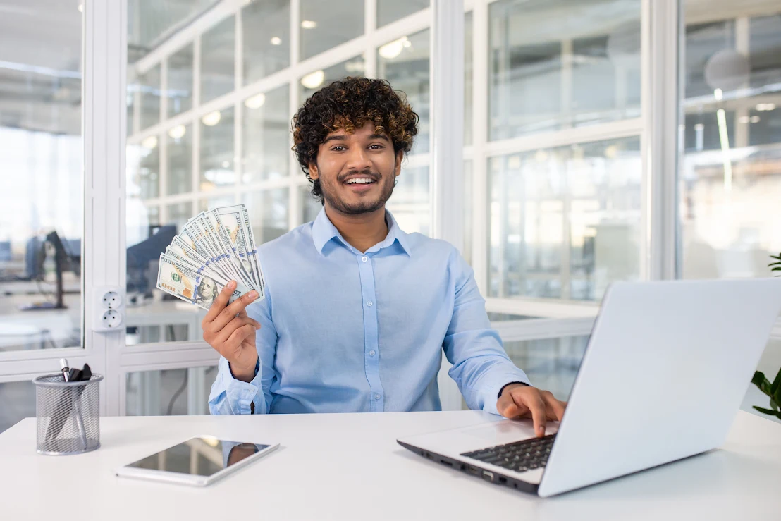 An employee smiles at his computer while holding a stack of money.